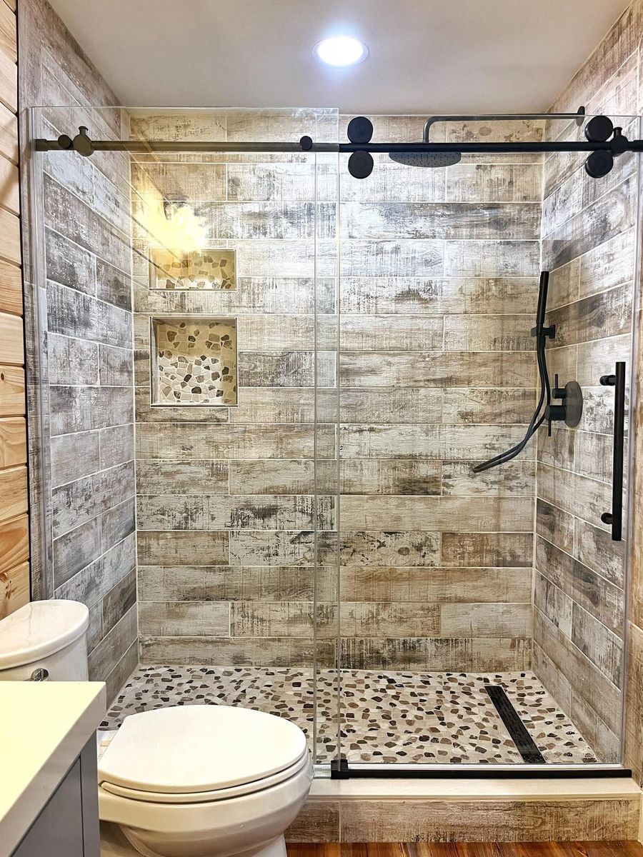 Custom shower with wood-look tile, pebble floor inlay, and a sliding glass barn-style door with matte-black hardware